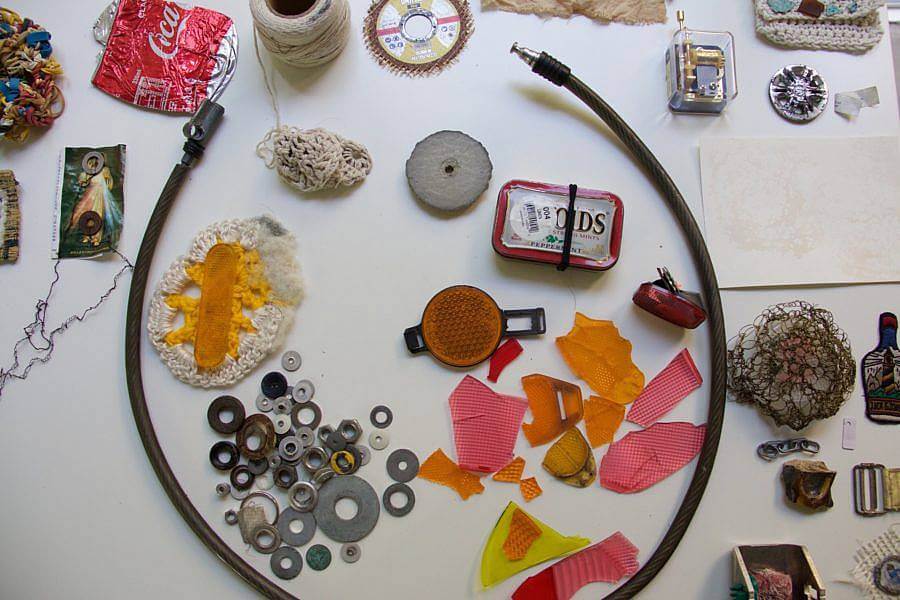 Image Description: pictured here is a small photograph of Dalbey's work space. Spread out on a desk are some metal washers, found shards of plastic, cables, twine, and paper.