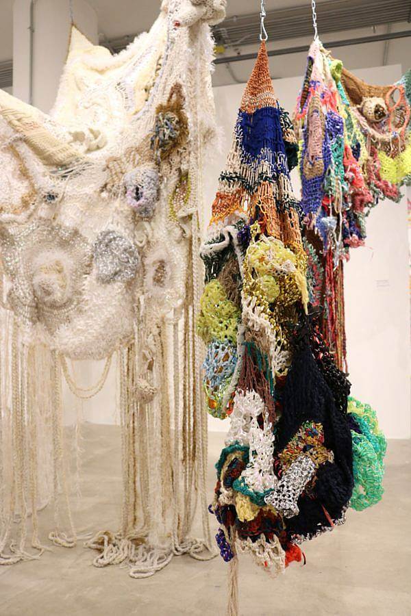 Image Description: In this photograph Dalbey's work is being exhibited. Two very large knitted masses are being hung from hooks and chains attached to the ceiling. The piece on the left is beige and ivory and much of it hangs from the suspended mass and touches the floor. The piece on the right is multi colored and more intricate, it hangs nearer to the floor that the piece on the left.