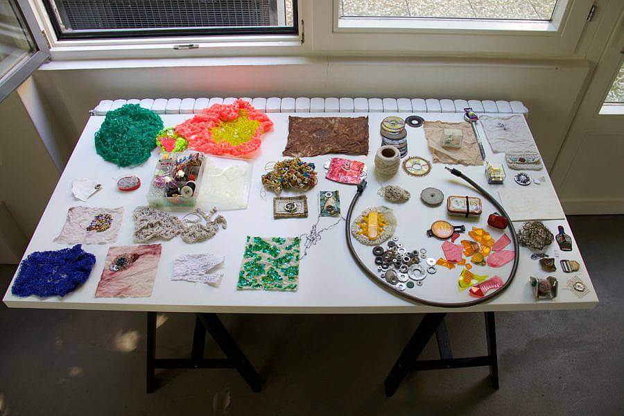 Image Description: This image is a larger photograph of Dalbey's table in her home studio. This photograph features the same plastic, washers, and cables, in addition to other colorful knitted and woven masses. Other small sewing materials are also featured.