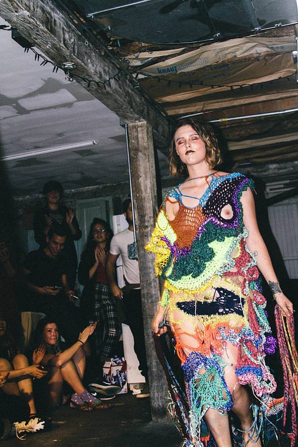 Image Description: This photograph features a model adorned in Dalbey's work. They are walking at an event amongst an audience, their garment features colorful, loosely knitted sections of yarn that cover the model from their shoulders to their ankles. 