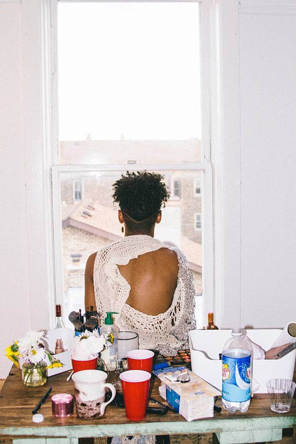 Image Description: In this photograph a model is wearing one of Dalbey's knitted works. The model is facing away from the camera and is looking out of the window that is in front of them. The knitted work is ivory and has a large hole that exposes the model's back and shoulder. The model is in a domestic setting with drinks and household items on a table between them and the camera.