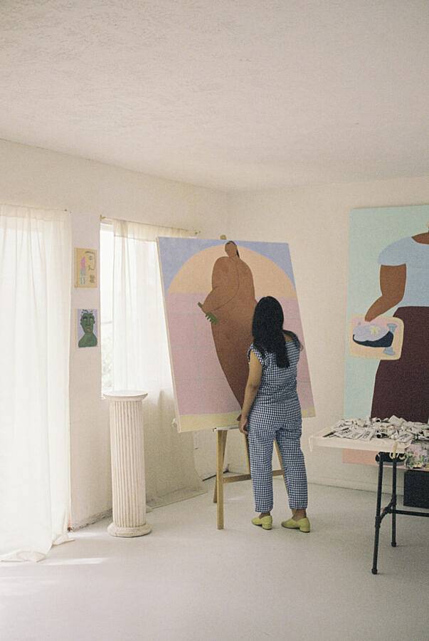 Image Description: This photograph features Martinez working in her studio. Martinez is working on a large painting on an easel accompanied by her other works in a white room with sun shining through the drapes. Martinez is wearing a plaid pair of pants and matching top paired with green shoes.