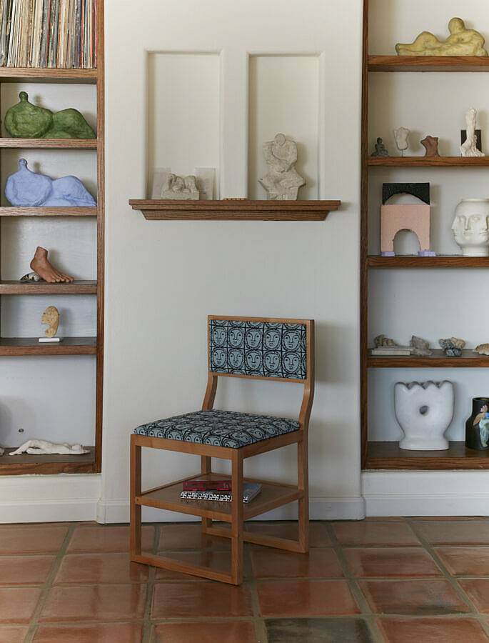 Image description: This photograph features a furnished  domestic scene. The walls are white and the floor is made up of glossy brown tiles, a single wooden chair with blue sun upholstery is set between shelves of ceramic objects and vinyl records.