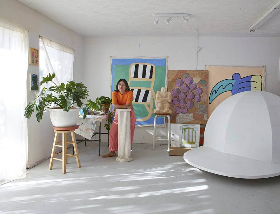 Image description: This image features Martinez in her studio with some of her work. On the wall behind her are un-stretched canvases, a large sculpture resembling a baseball cap, a pillar that Martinez is leaning against, and a houseplant on a stool.