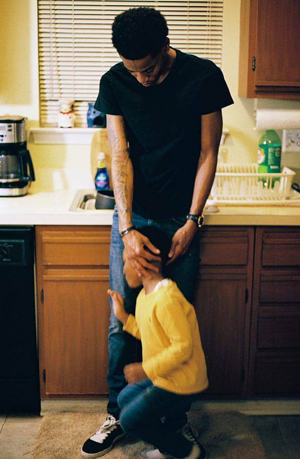 Image Description: This is a photograph of a father in a black t-shirt standing in front of a kitchen sink. There is a young child in yellow in front of him and the father is reaching down to try and calm him to be in the picture.