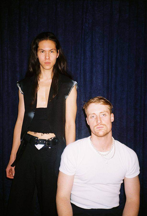 Portrait of Steve Katona and Billy Bultheel in front of dark blue curtain. Billy wears a white t-shirt and to his left,Steve wears a black vest and black wrap around their stomach.