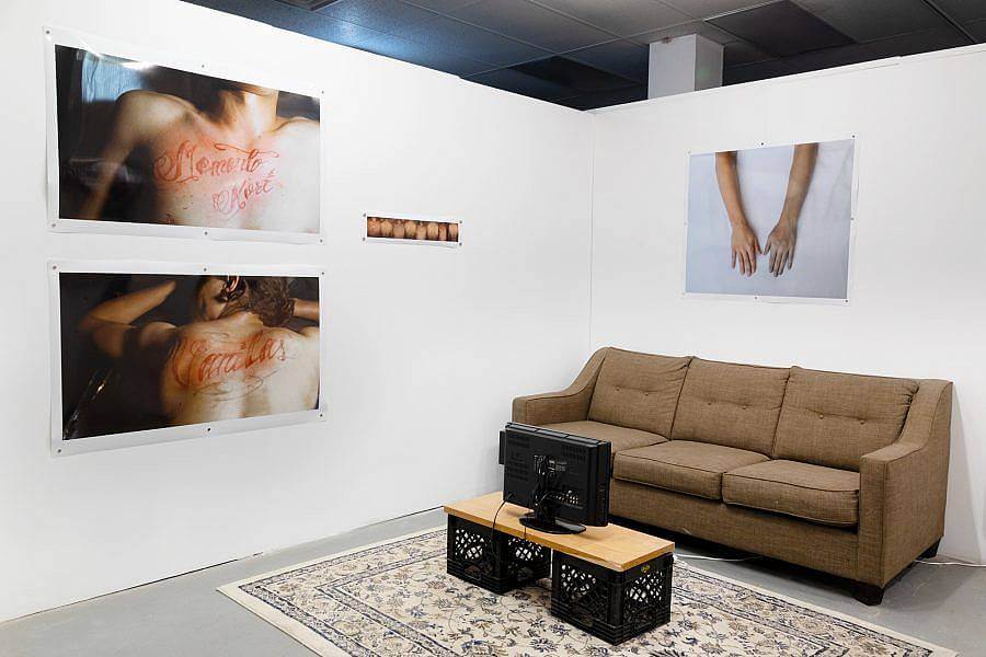 The corner of an exhibition is on display. There are two white walls, against the right wall is a tan mid century couch. On the ground a repurposed oriental rug. Atop the run is a small old flat-screen tv is on a wood plank held up by milk crates. On the Right wall is a photography of outreached hands. On the left are two large photographs of tattoos in red ink on someones chest and back. Next to this is a photostrip. 