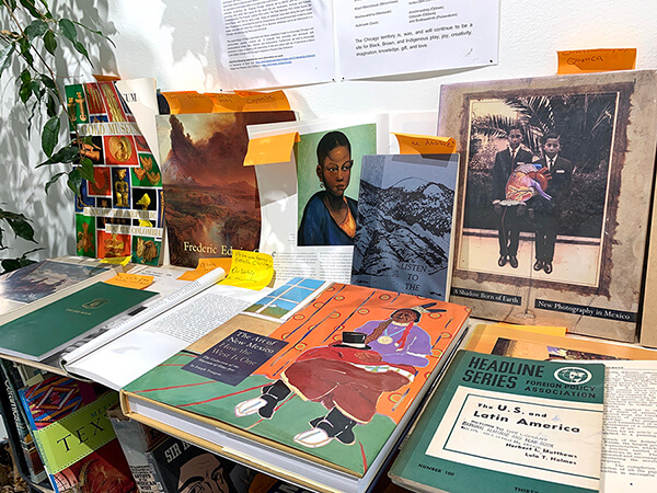 A variety of books of all different coors, several of them green have orange sticky notes sticking out from them. Several of the books are turned open to the sctions that are sticky-noted. The center book features an illustrated woman with black hair and a strong forehead. Behind her is a light green background. 