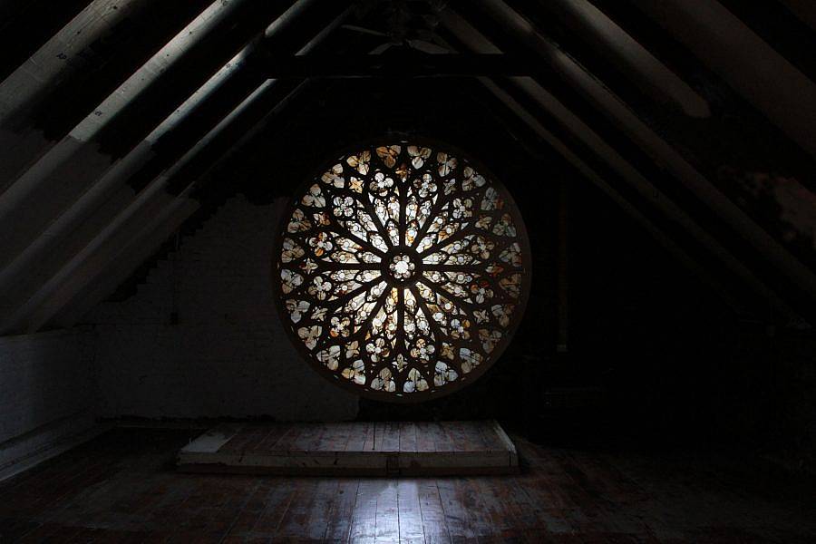 View of a circular "Glass paned window," formed by dried lettuce. The window is at the back of a attic space. Light shines through it. 