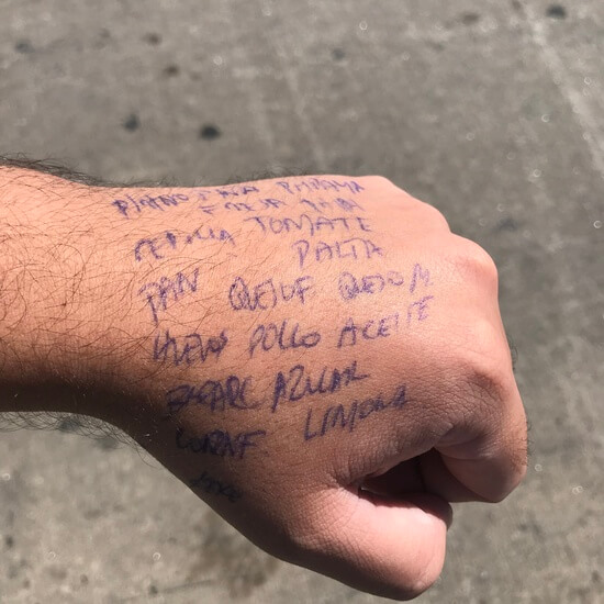 Photo of artist's hand above a grey cement pavement. There is blue lettering on the hand. It is a grocery list in spanish. The visible words going from left to right read as 'PINTO,TOMATE, PAN,PALTA, QUESO F, QUESO M, HUEVOS, POLLO, AZUCAR, LIMON." There are other words as well, they are not all readable. 