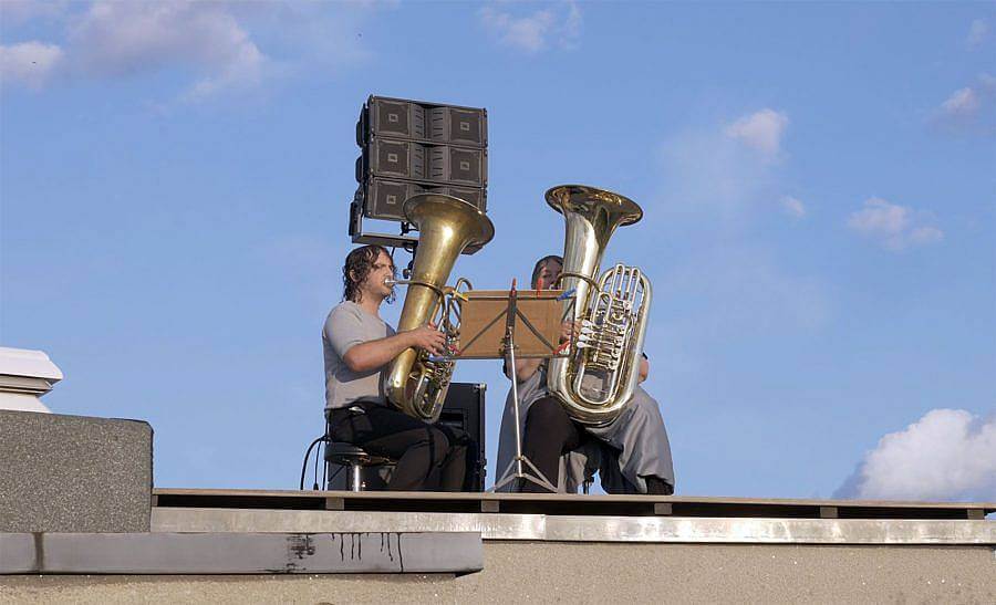 Photograph of two tuba players on roof top of a building. A blue sky with sparse clouds in visible behind them. The sun shines on their face..