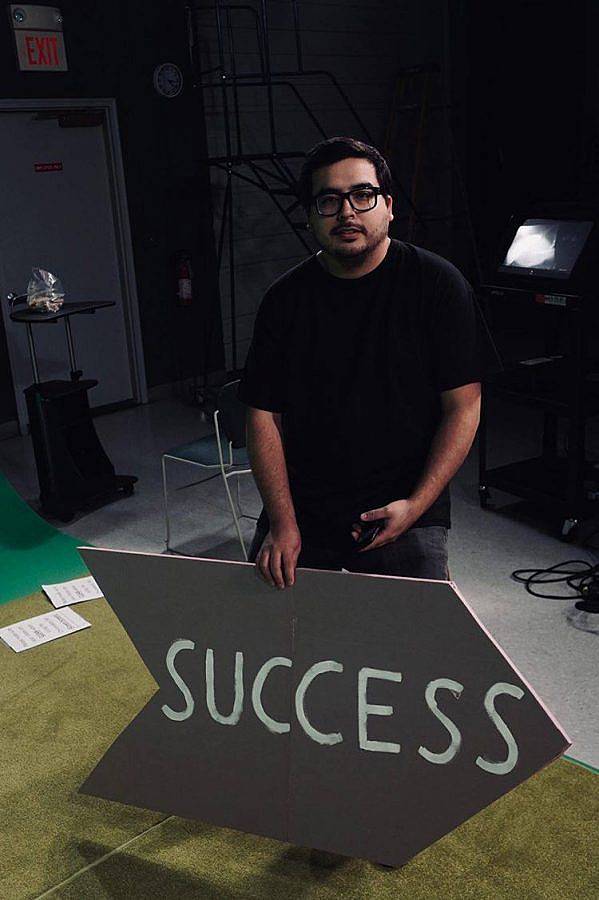 Colored photography of artist Gonzalo Hernandez. He is pictured in a black shirt with black glasses on a green carpet near a green screen. He holds a large arrow sign that reads "SUCCESS."