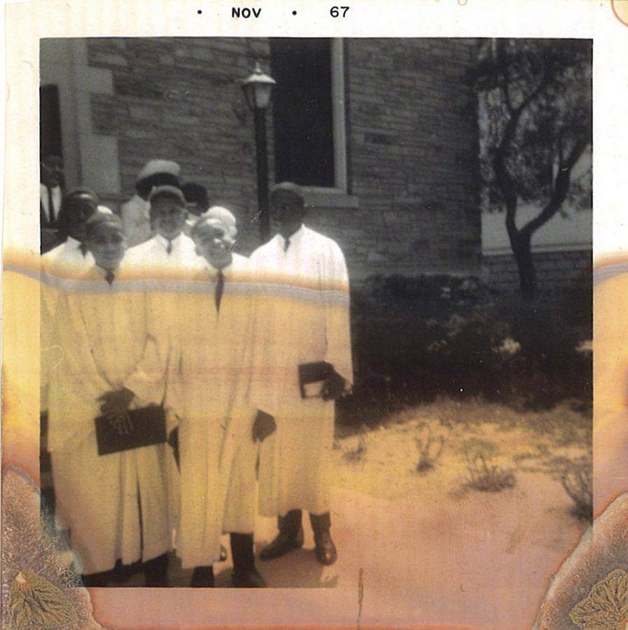 Image Description: This is a photograph from November of 1967. There is a group of five young boys in white robes for their confirmations standing to the left of the image. They are standing in a garden with a stone building and a lamp post behind them. The photograph itself is deteriorating.  