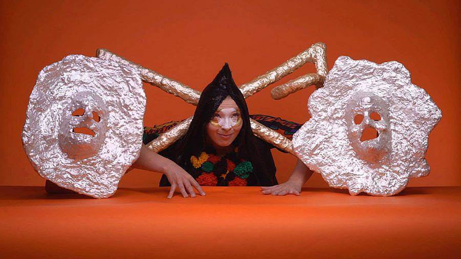 Performer Emili Losier places their hands atop an orange surface, in front of the same orange background. Their hair is fashioned to a paint. White circle is painted in the middle of their face. A chromatic and shiny sculpture is attached at the neck, which contains tubes and cast human faces.