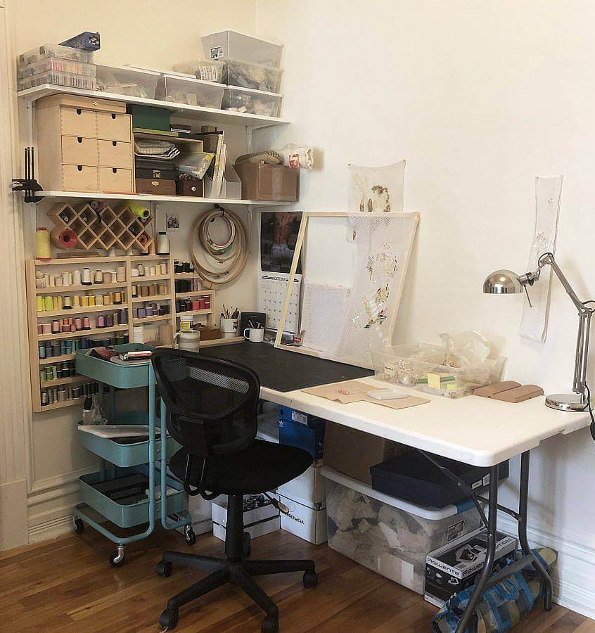 Studio of the artist depicted. Shelves include spindles of thread, boxes of sequins and more. A desk and chair is also depicted. 