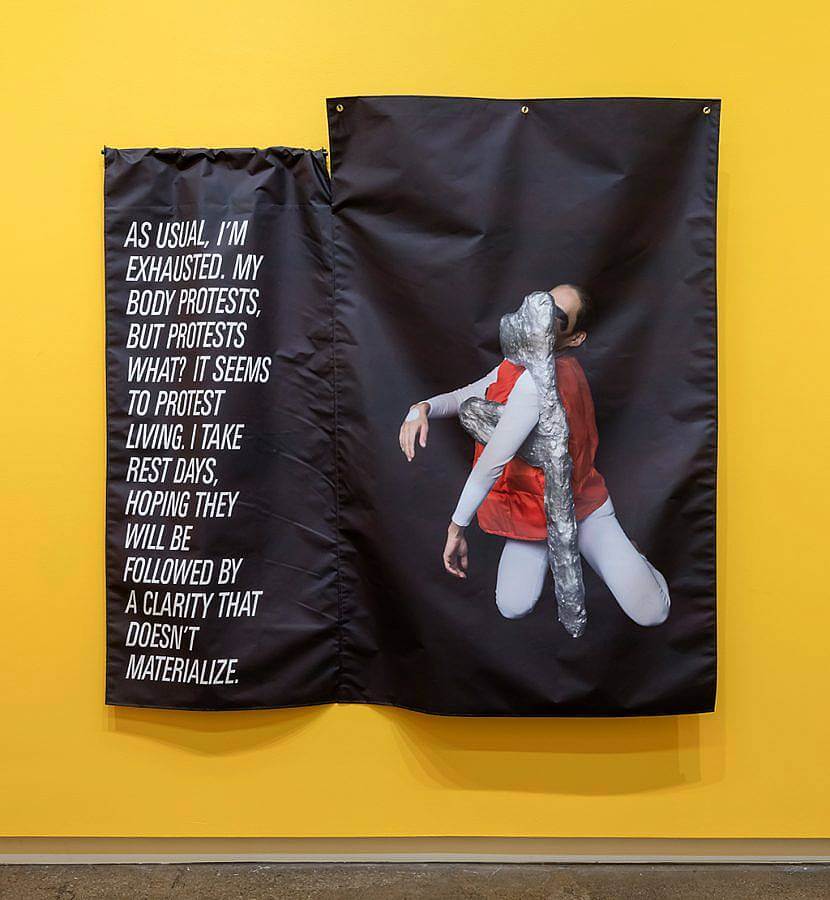 A black poster is hung against a yellow wall, on the right is a text message, "As usual, I'm exhausted. My body protests, but protests what? It seems to protest living. I take rest days, hoping they will be followed by A clarity that doesn't materialize." To the right a performer moves with a silver hand made sculpture, their arm extends over part of the sculptural object.