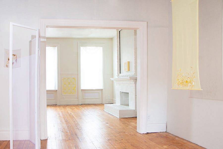 Installation view of Indecent Exposure. A yellow translucent partition hangs from the ceiling, various works are visible and and installed along the walls. An arch way is depicted which leads to another room and fireplace. 