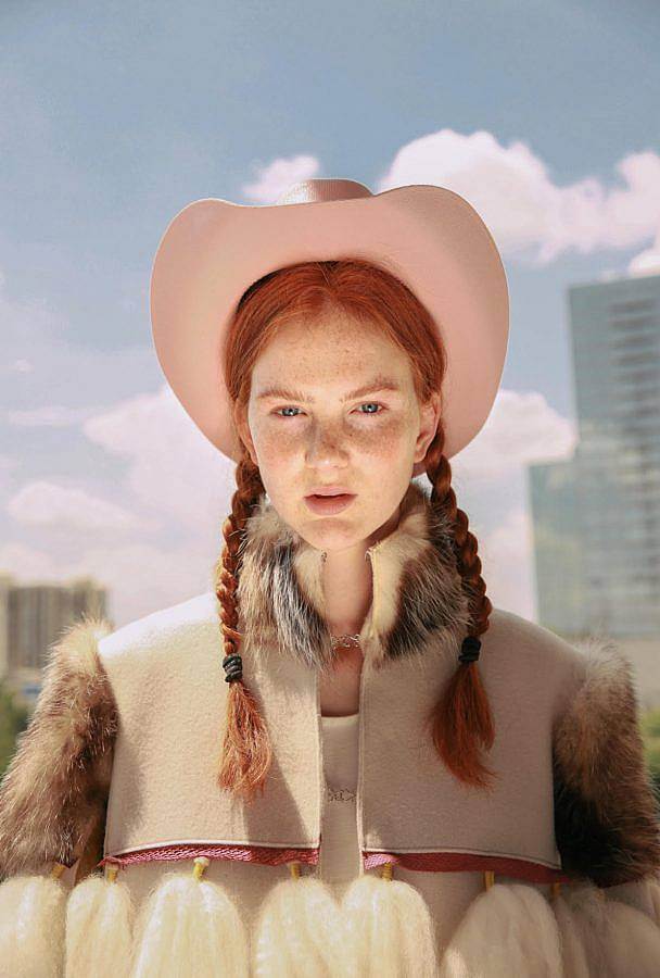 Photograph of a red-haired white feminine person outside. They wear a pastel pink cowboy hat, their hair is in braids, and they have on a fur coat, which has a fur color, shoulders, and the rest is a suede material. There are also white tassels of brushed out hair visible. Behind them are sky scrapers and a partly cloudy sky. 