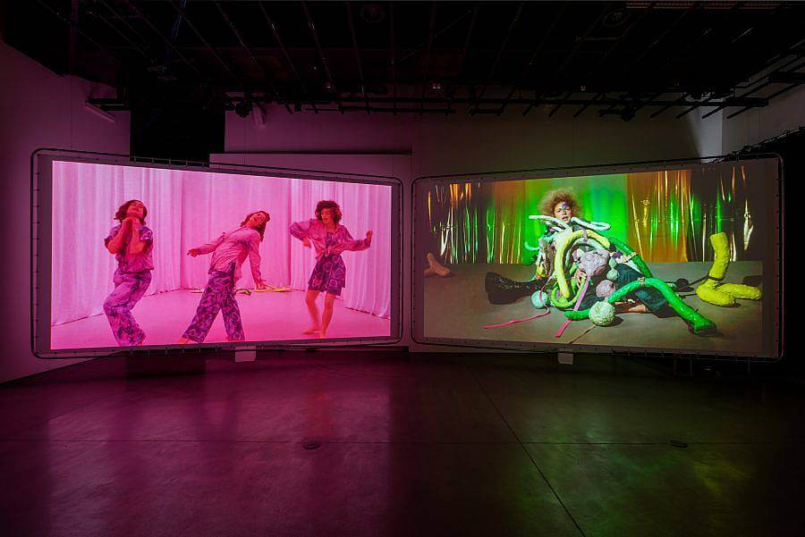 Image of two-way video installation. On the left a video of three performers in a pink curtain background dance. In the left screen is a performer, a gold curtain background is behind them. A plethora of objects are placed upon them and are apart of them. Green lights are used in this video.