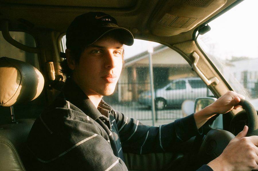 Dan wears a black baseball cap and a stripped polo long-sleeve shirt while driving in the front seat of a Toyota. The sun shines through the front window onto Dan's face.