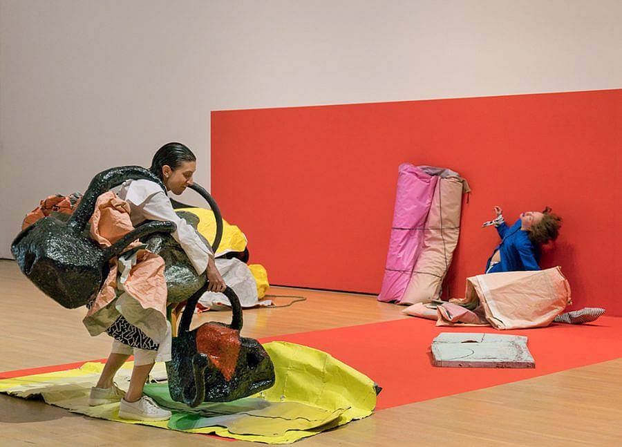 Performance with two performers, red background on wall, and orange rectangle are on floor. In foreground, one performer with their hair slicked back has multiple bag-like made objects on their body/apart of their body. They pick up another. In the background to the right is the second performer, their back extended backwards and they are wrapped in fabric against the red wall.