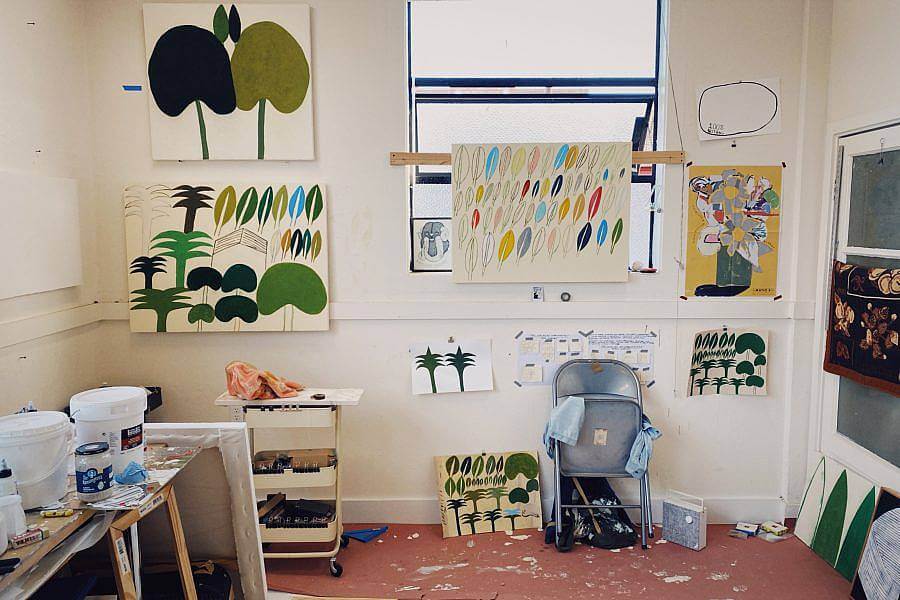 Photograph of Ryan Whelan's studio, features paintings of his hung on the walls, painting supplies and a metal folding chair.