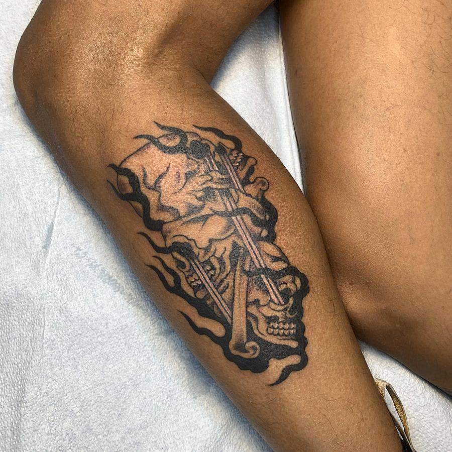 Photo of a tattoo on a calf. On the brown skin is tattoo of three skulls atop one another. Swords come out of their eyes. Black flame linework encircles the skulls.