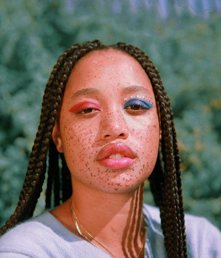 Photograph of a black woman, with braids and freckled skin in the sunlight. Her left eye is painted red, her right blue, with stars above it. Her hair is done in a box braided style. Green bushes and foliage are visible behind her. 