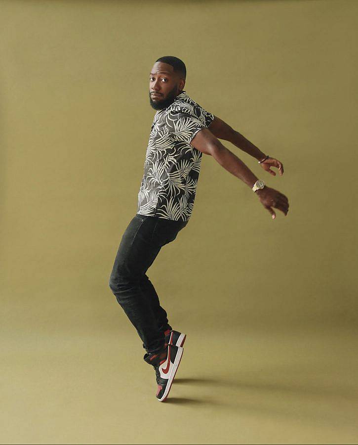 Editorial photography of actor Lamorne. Photo of a black man in a toe stance against an olive background. He wears nike shoes and a patterned top. He has a trim beard and his hair is cut close to his head.