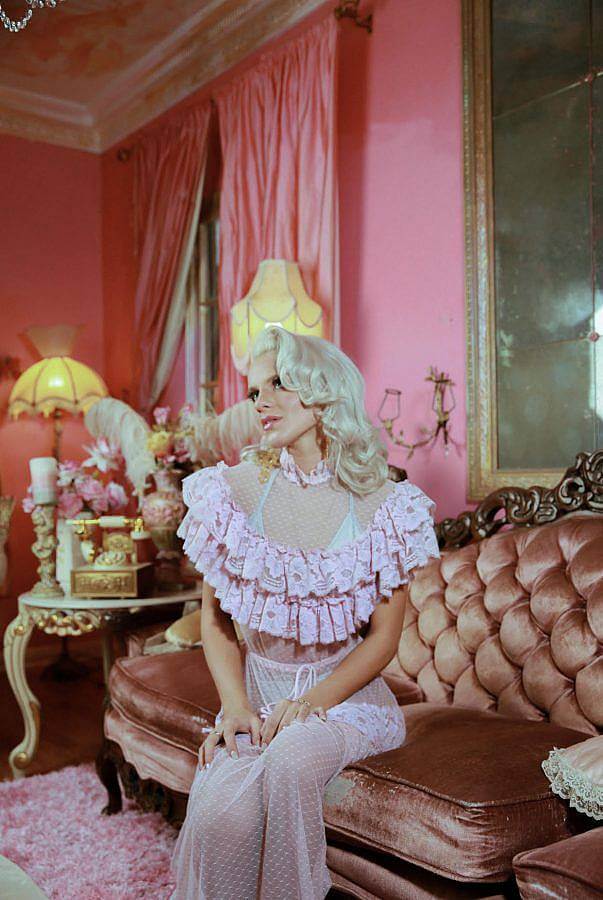 Inside photography of a feminine white person with platinum blonde shoulder length hair. They wear a pink ruffled transparent dress. Beneath it they wear a white bikini top and bottom. They sit on a rose gold antique couch, and opulent shades of pink curtains, walls, flowers and gold lamps and tables are behind them. 