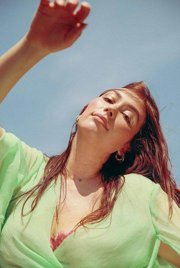 Photo of a woman with warm olive skin and long warm brown hair. The photo is take from below and the subject smiles, with her hand in the air. A clear blue sky is behind her. She wears a soft lime green translucent top and a mauve lace bra.