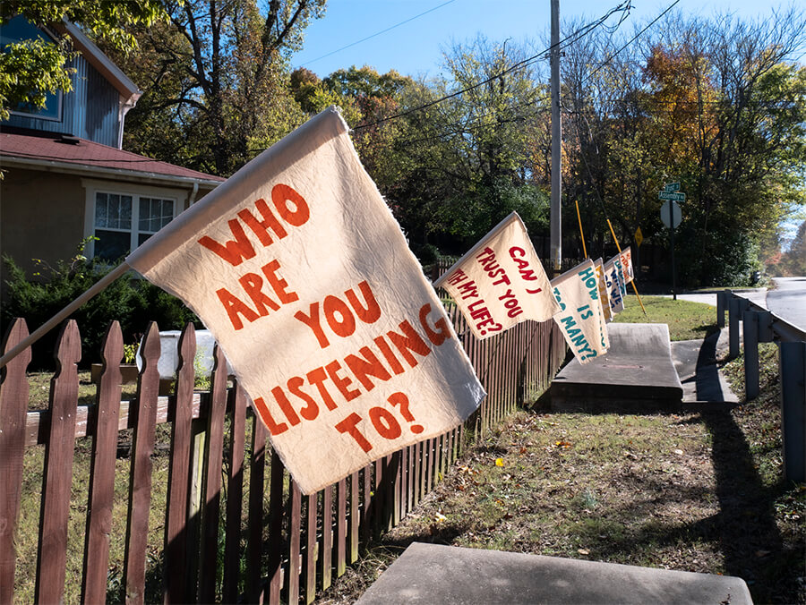 A series of flags are hung off of a wooden Fence. The one closest to the viewer reads, "Who are you listening to?" , The other about 20 ft back reads "Can I Trust You with My Life?" There is grass around the fence, trees in the background, and a blue sky.