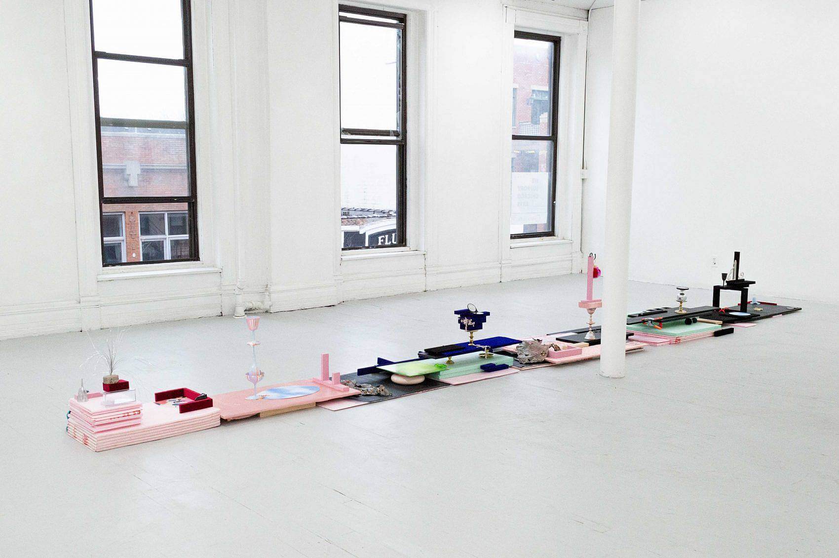 There is a long, low, and narrow sculpture sitting on the floor. There are various stacks of of light pink, green, and black insulation foam topped with various small objects like tinsel, small blue towels, compact mirrors, rhinestones, or small makeup sponges. There are also many small velvet blocks in blue and red.