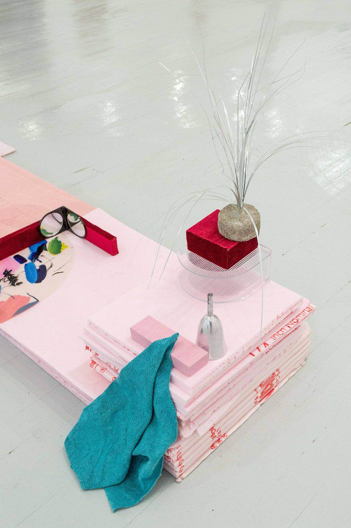 Leaning against against a stack of square thin sheets of pink foam is a blue rag pulled up to the top. There is also a transparent half-oval shaped napkin holder with a red velvet block on top. On top of the block is a short cylinder of cement with silver tinsel poking out of the top.