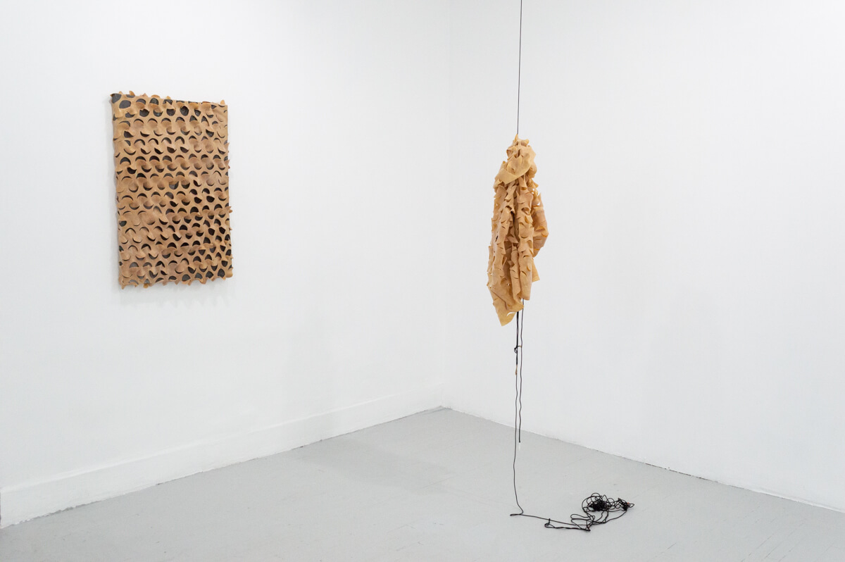 On the left side of the corner, a black rectangular canvas wrapped in tan latex with a grid of half-circle cut-outs. On the right is the same latex material hanging from black paracord further out into the space.