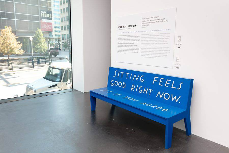 Image description: A blue bench that says, "Sitting feels good right now. Sit if you agree." Behind it is wall text with the heading “Shannon Finnegan.” To the side is a window looking out on downtown Denver.