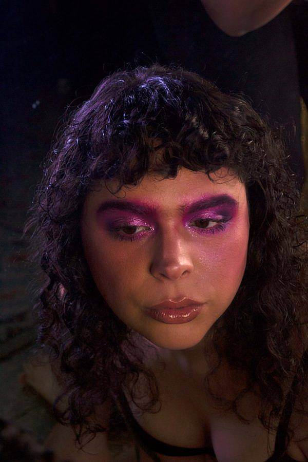 Photo of DJ Ariel Zetina, an olive skinned woman with curly dark brown/nearly black hair. It is a darkly lit photo with a spotlight on her hair and face. She wears saturated purple and pink make up on her eyes and a light glistens off her curly hair. 