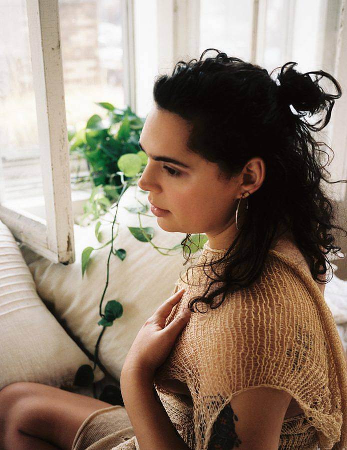 Photo of DJ Ariel Zetina, an olive skinned woman with curly dark brown/nearly black hair. She wears a streched tan mesh material and sits next to an open window and philodendron. 