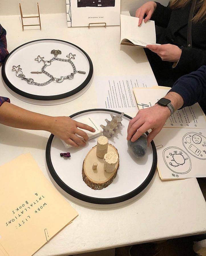 Image description. Hands holding and reaching for small artworks displayed on two Oxo-brand lazy susans. The objects include a deck of cards, a rock-shaped object, a 3D print of vertebrae, and some pill-bottle-shaped mushroom material resting on a slice of wood.