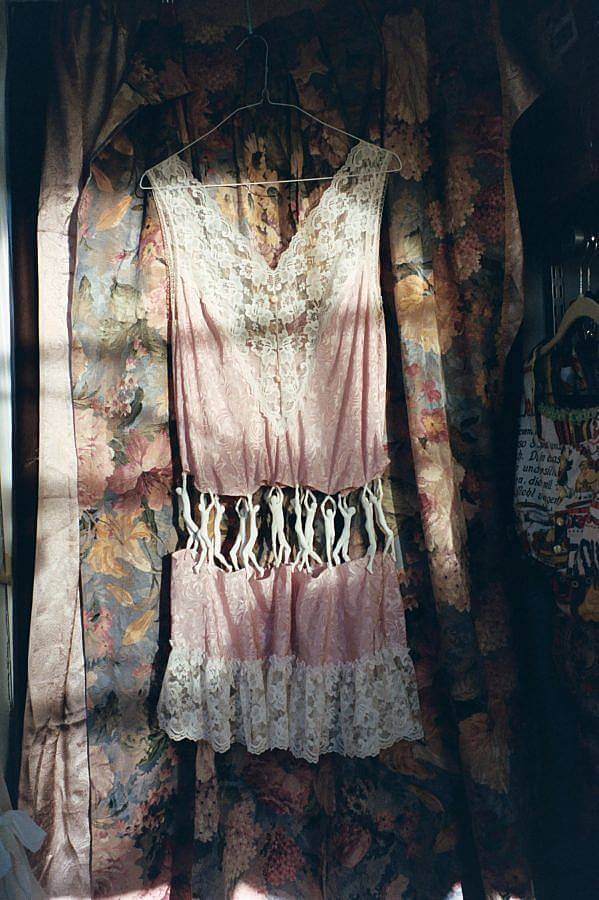 Image description: A lace lingerie dress is photographed in natural light against a vintage flower tapestry. In the middle of the dress a series of plastic clinging men figures hold the piece together. 