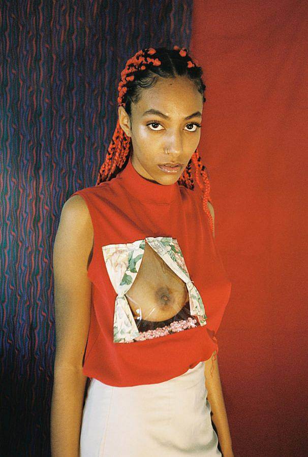 Image description: Model Awa Kaloga, a black woman with long red braids is pictures wearing a red turtle nexk top. Over her right breast a window pane reveals her chest. 