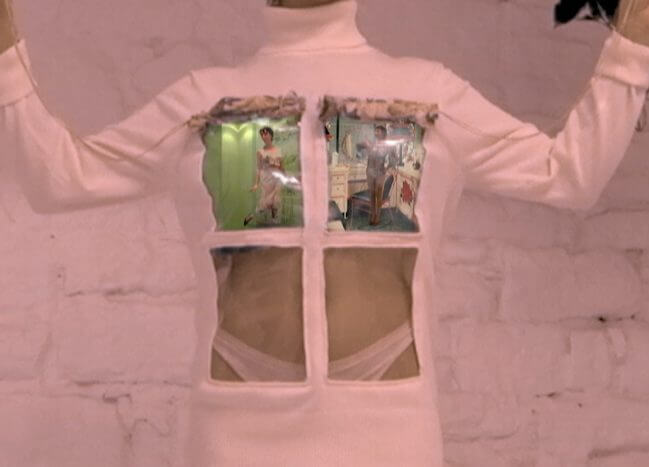 Image description: A still from Martina Cox's ad campaign. A model with pale skin wears a turtleneck dress with a window pane in front. In the top two panes of the window a series of video displays project.