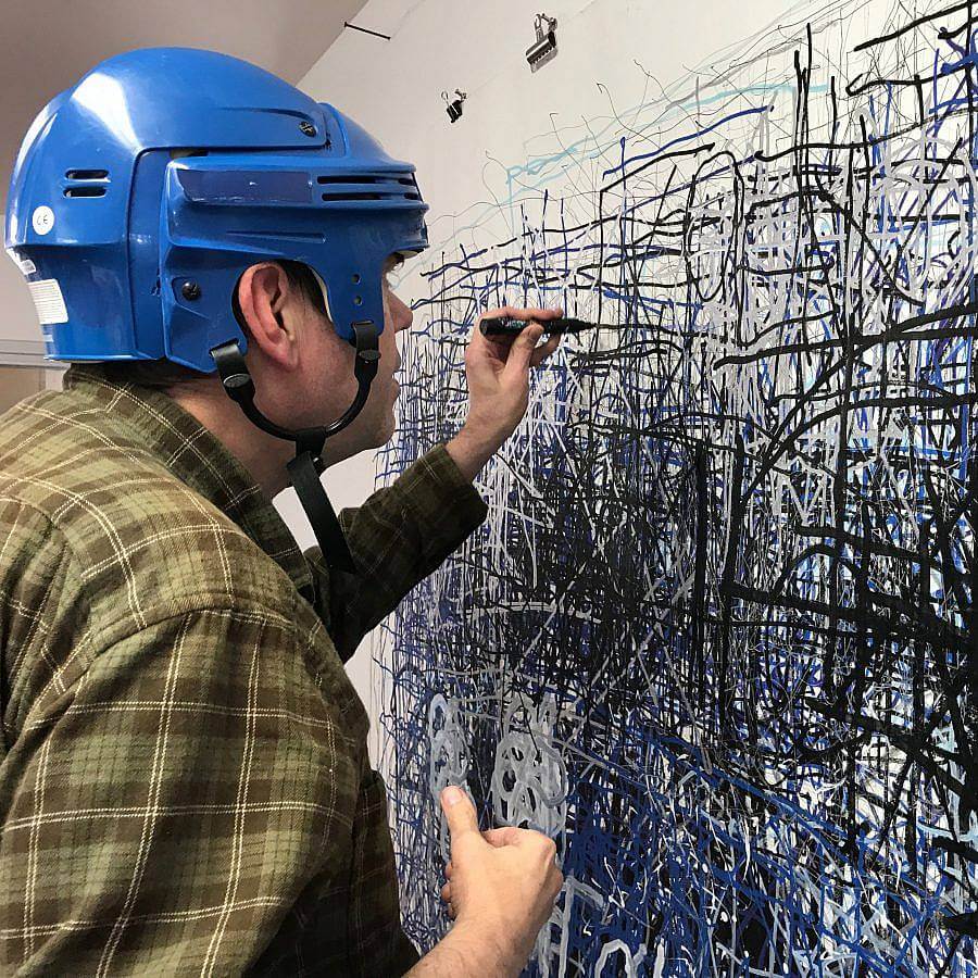 Image description: Artist Dan Miller wears a blue protect helmet and an olive green plaid shirt. He is using a black marker to make marks on a large white surface. We can see the profile of his face.