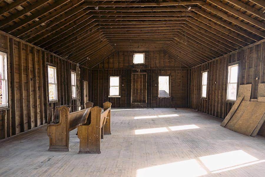 Image description: A stripped down all wooden interior of a building is displayed. Natural light pour in through the windows. Two church pews are on the left.