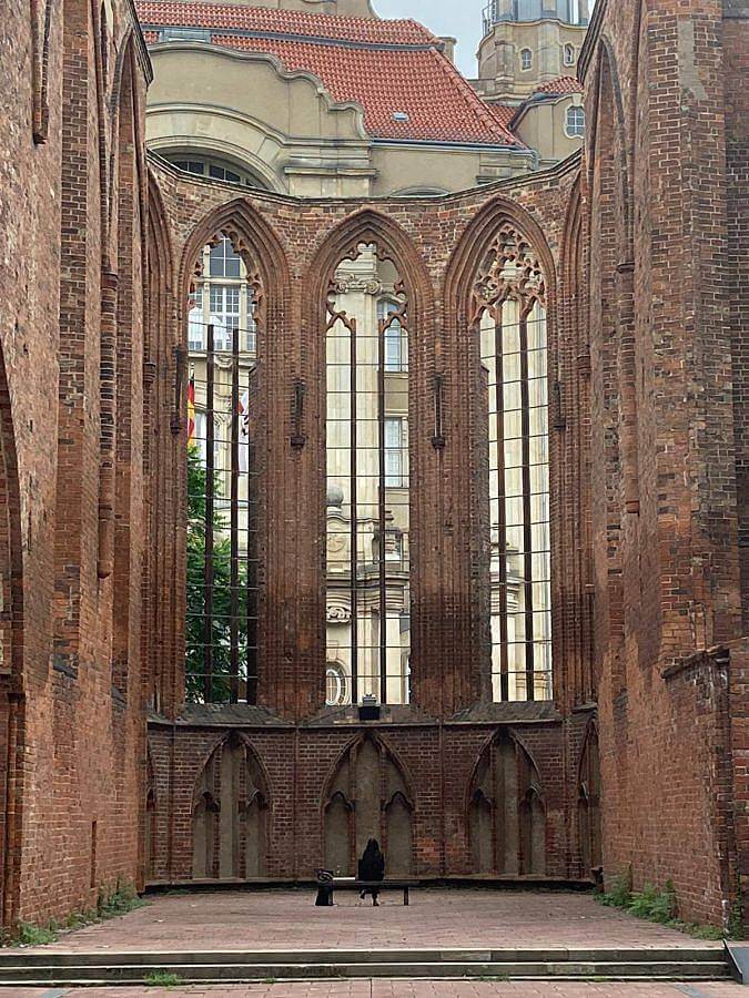 Image description: An individual sits on a bench inside a massive church ruin courtyard. Three arches windows frame the structure. 