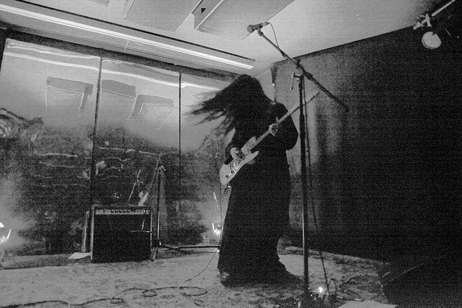 Image description: Black and white photograph of Artist Johanna Hedva in front of three reflective panels. Their long black hair whips around them. Their light guitar stands out against their black outfit. 