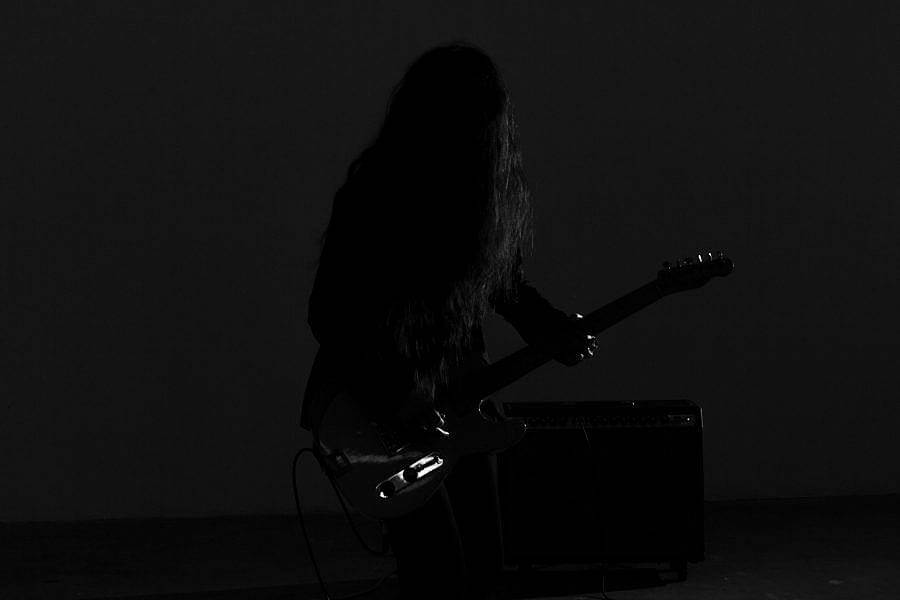 Image description: Stylized dark Black and white image of artist Johanna Hedva with their guitar. Their long black hair is in front of their face. Highlights of light on the guitar are visible. The artist is subtlely visible against a black background.
