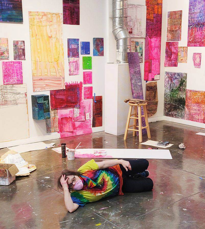 Image description: Artist Nicole Storm lays on the floor with a light pink mask over her face. Her colorful artwork covers the walls on both sides. There is pink coloration on the walls painted or drawn on.