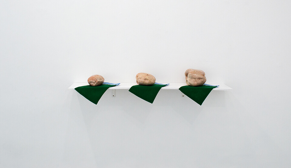 Image Description: There are three pink-toned tan amorphous ceramic pieces on a white shelf. They are evenly spaced and each rests on a dark green mat with a light blue stripe.