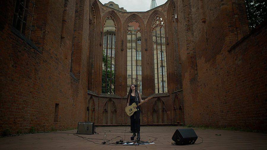 Image description: Artist Johanna Hedva performs in amidst the courtyard of church ruins. They are in the center of the courtyard with their guitar and two amps, one on each side of them.  